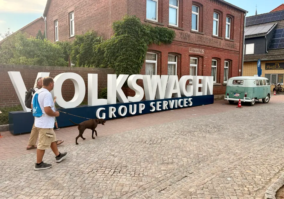 A couple and their dog walk past a large 3D VW Group Services logo with a VW Beetle Campervan in the background